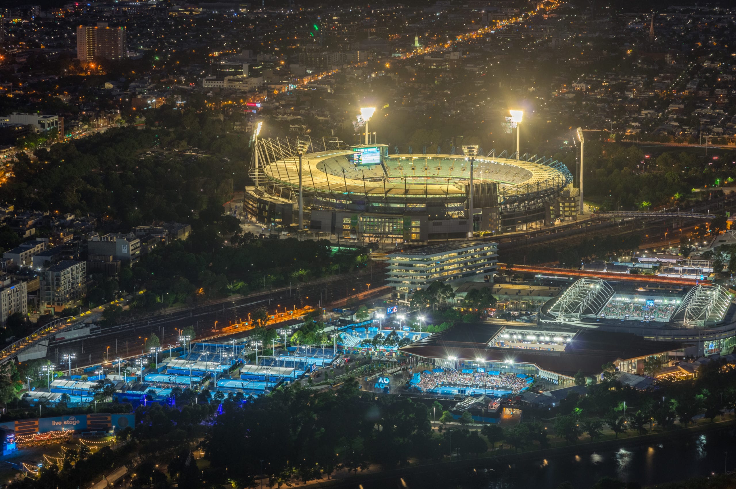 Melbourne Cricket Ground and Yarra Park tennis stadium illuminated at sunset.